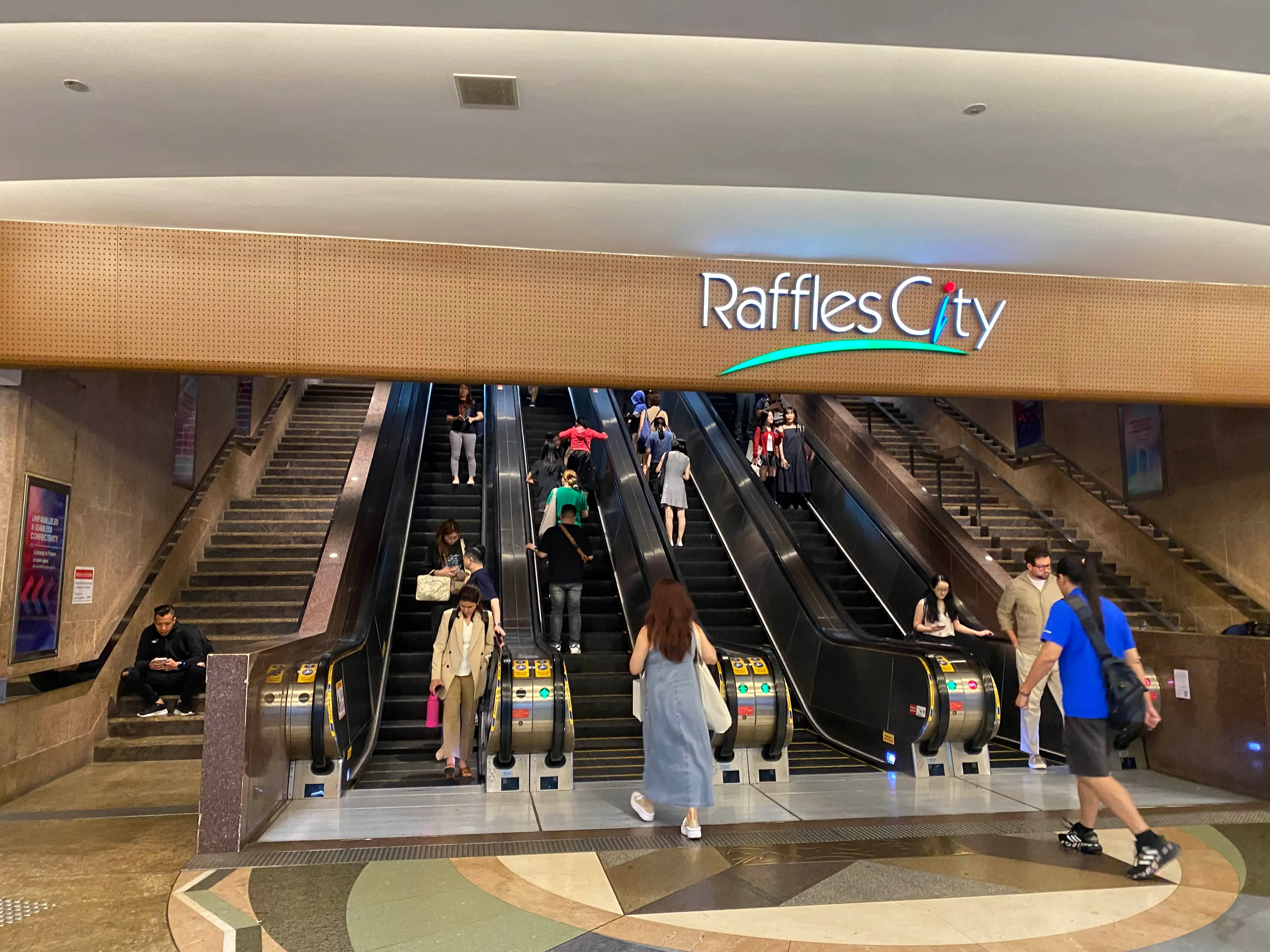 Exit A of City Hall MRT that leads to Raffles City mall.There are escalators and stairs that lead to street level. The Raffles City logo is displayed on the signage above the escalators.