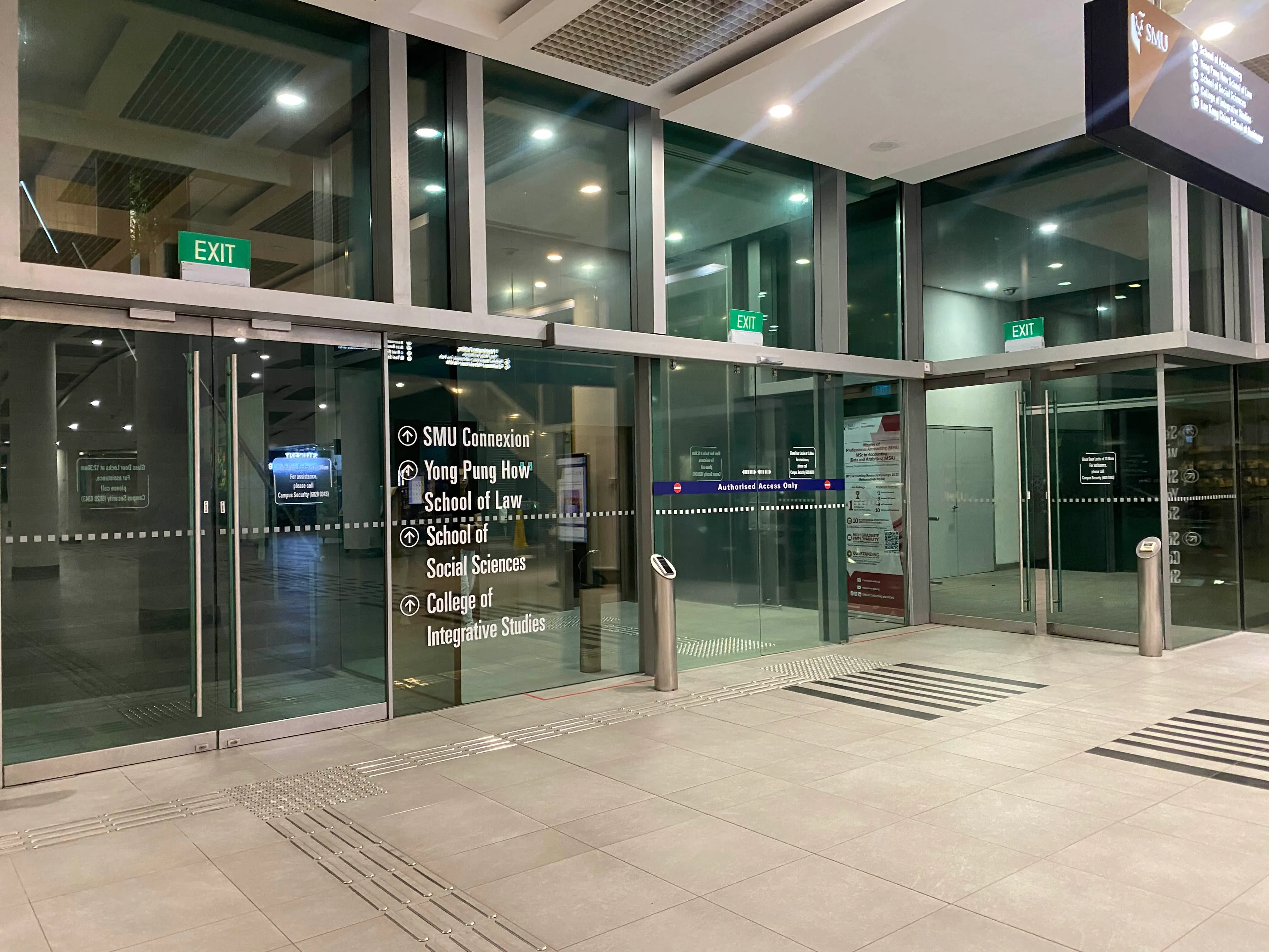 Several glass doors that lead to street level. The door on the left next to the wayfinding sign leads to a flight of stairs. The door in the middle leads to a lift that is gated by keycard access. The door on the right leads to an escalator.